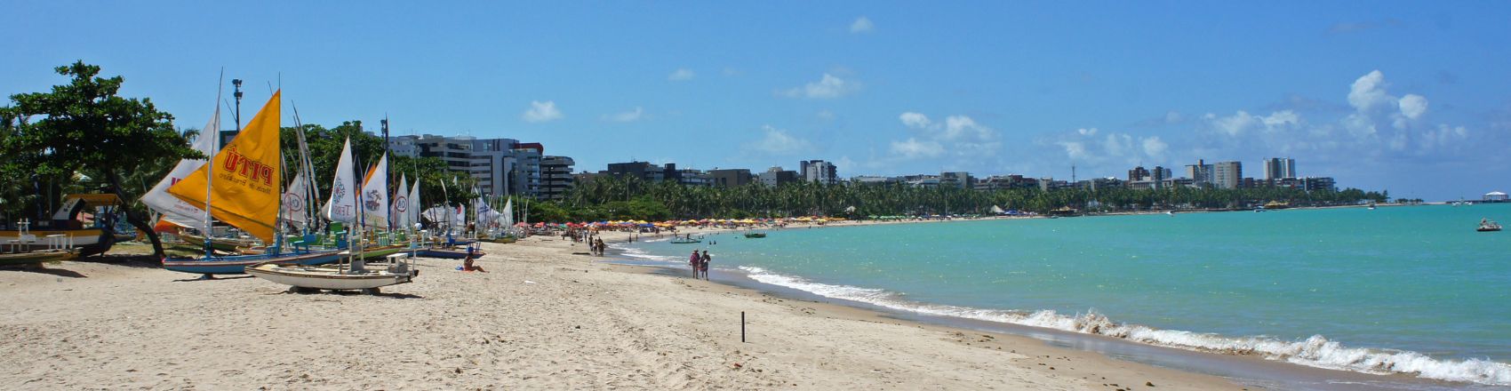 Vista da praia em Maceió. Há uma extensa faixa de areia com banhistas caminhando. Uma onda quebra do mar azul e calmo quebra na orla. Na areia há barcos ancorados e ao fundo, na linha do horizonte, prédios e vegetação.
