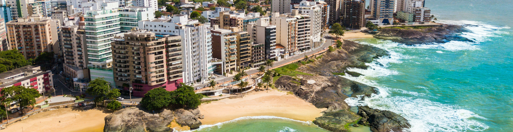 Vista aérea da orla da praia de Guarapari. o mar azul cria um constraste com as pedras e os prédios à sua frente.