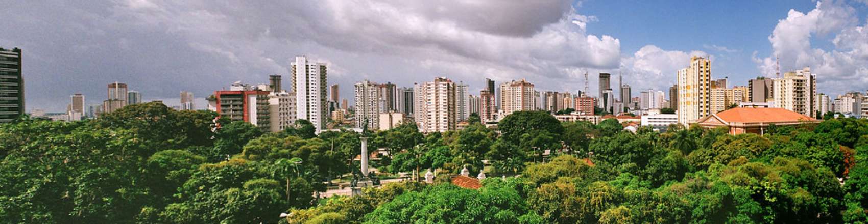 Na foto há árvores em primeiro plano, um monumento ao meio e prédios ao fundo cobrindo a linha do horizonte. O céu está nublado e com nuvens escuras.