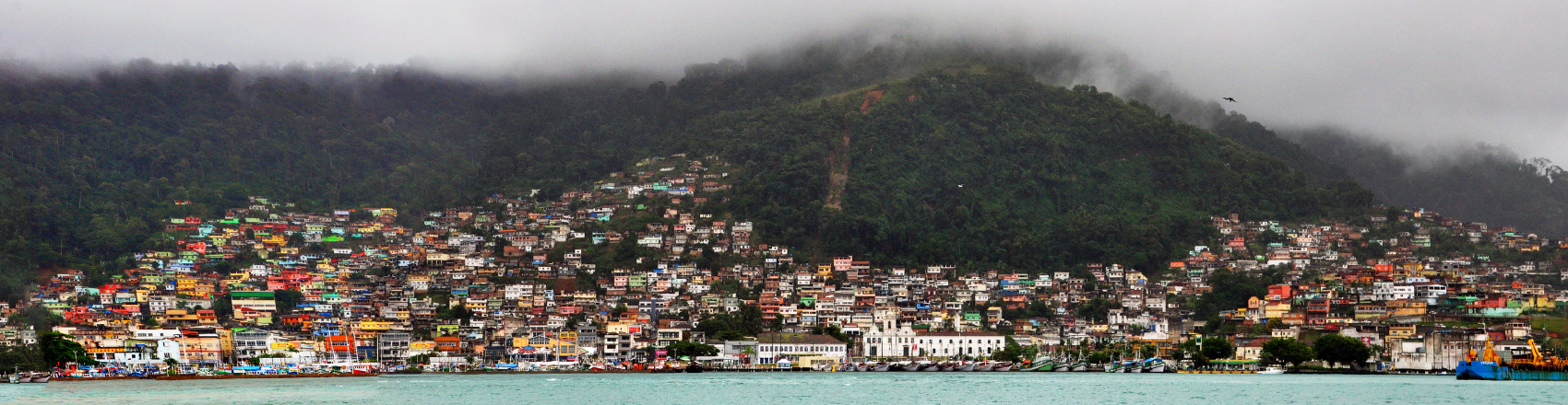 Vista panorâmica da cidade de Angra dos Reis. Há casas à beira-mar na frente e ao fundo montanhas com vegetação. A neblina cobre parte da paisagem