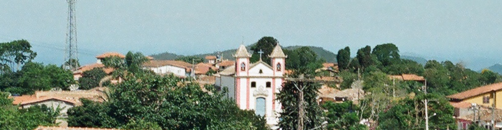 A imagem mostra uma paisagem com uma igreja no centro. A igreja possui duas torres com sino, detalhes em rosa e uma cruz no meio. Ao redor há casas e ao fundo montanhas.