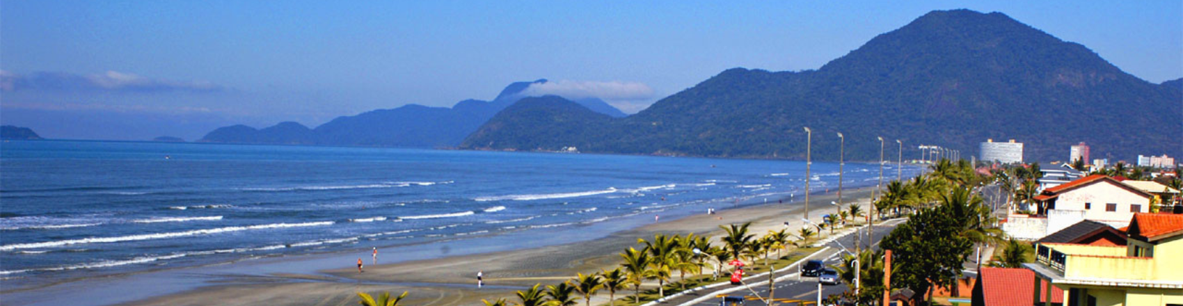 Orla da praia de Peruíbe, com mar azul, céu limpo e montanhas ao fundo. Ao atravessar a orla, há asfalto e casas, e pessoas andando pela areia e rua. 
