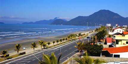 Orla da praia de Peruíbe, com mar azul, céu limpo e montanhas ao fundo. Ao atravessar a orla, há asfalto e casas, e pessoas andando pela areia e rua. 