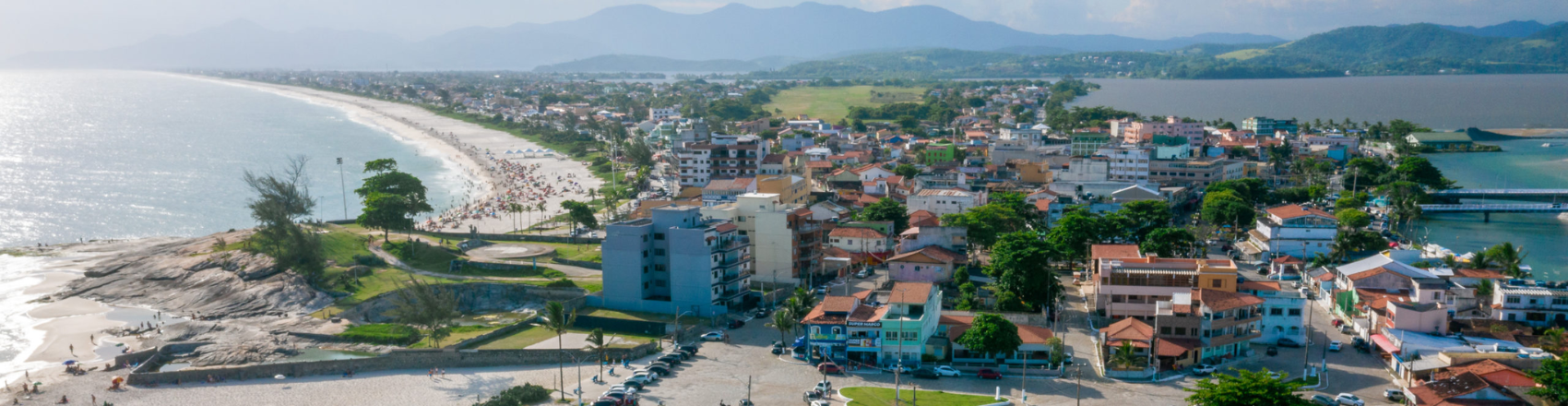 Na foto, vemos Saquarema, no Rio de Janeiro, com uma praia de um lado e uma faixa de terra separando o mar do outro lado. Nessa faixa de terra, vemos edifícios e árvores, e na orla da praia, temos pessoas e banhistas.