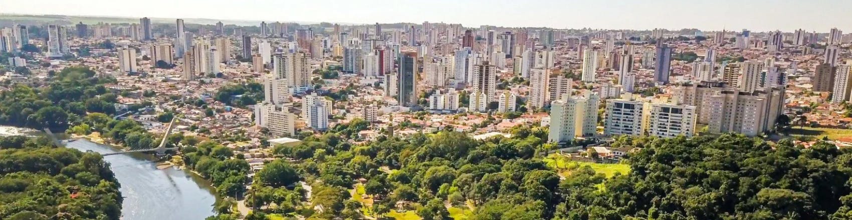 Foto aérea de Piracicaba, no interior de São Paulo, mostrando prédios ao fundo e um parque com muitas árvores de copas verdes e um lago cortando a cidade. 