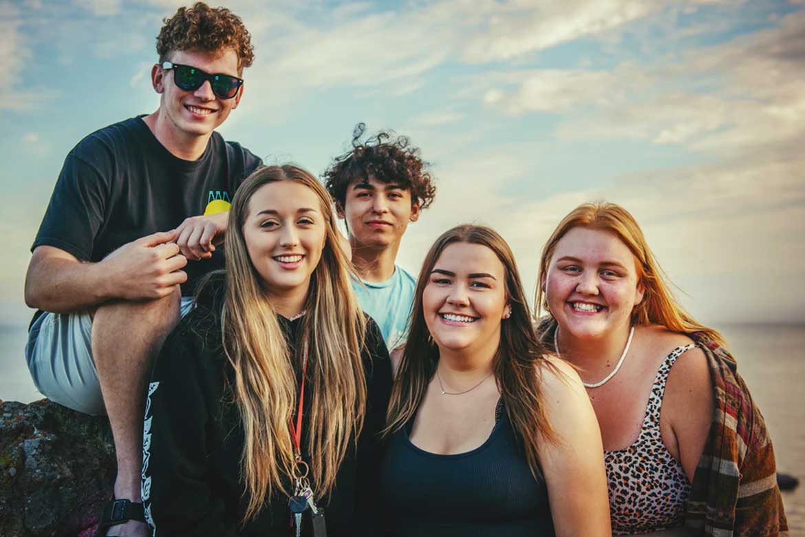 Grupo de amigos sorri em frente à praia.