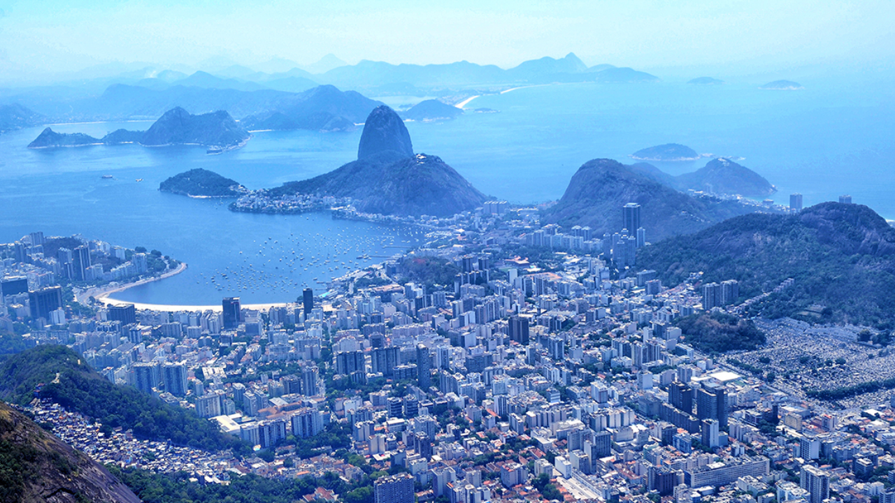 Visão panorâmica do Rio de Janeiro, com seus prédios em frente à orla e o Morro do Pão de Açúcar ao fundo.