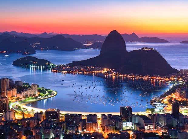 Vista panorâmica do Morro do Pão de Açúcar à noite, com o céu alaranjado e as luzes da cidade iluminando a orla da praia.