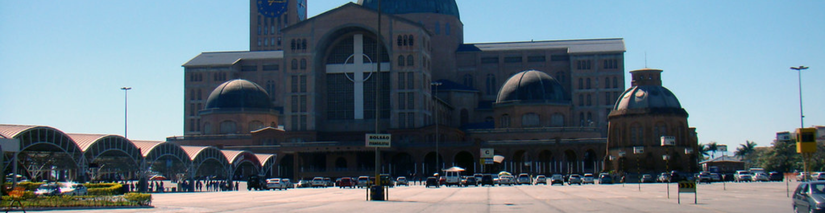 Fachada da parte da frente do Santuário de Nossa Senhora em Aparecida na Cidade de São Paulo. Na foto vemos os carros estacionados e no lado esquerda, temos uma estrutra em abóbada que cobre parte do pátio.