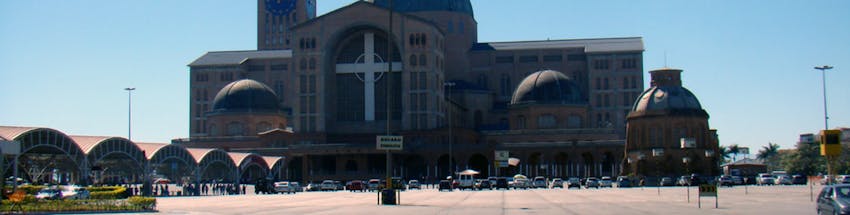 Fachada da parte da frente do Santuário de Nossa Senhora em Aparecida na Cidade de São Paulo. Na foto vemos os carros estacionados e no lado esquerda, temos uma estrutra em abóbada que cobre parte do pátio.
