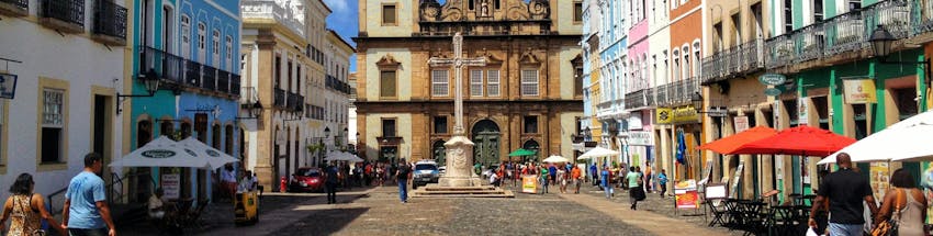Pelourinho em Salvador na Bahia. Na foto é possível ver a cruz da Igreja se São Francisco ao centro. Ao lado, além de guarda-sóis abertos, temos pessoas caminhando.