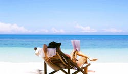 Imagem de mulher de costas, sentada em espreguiçadeira na praia, enquanto lê um livro no sol, com o mar à sua vista.