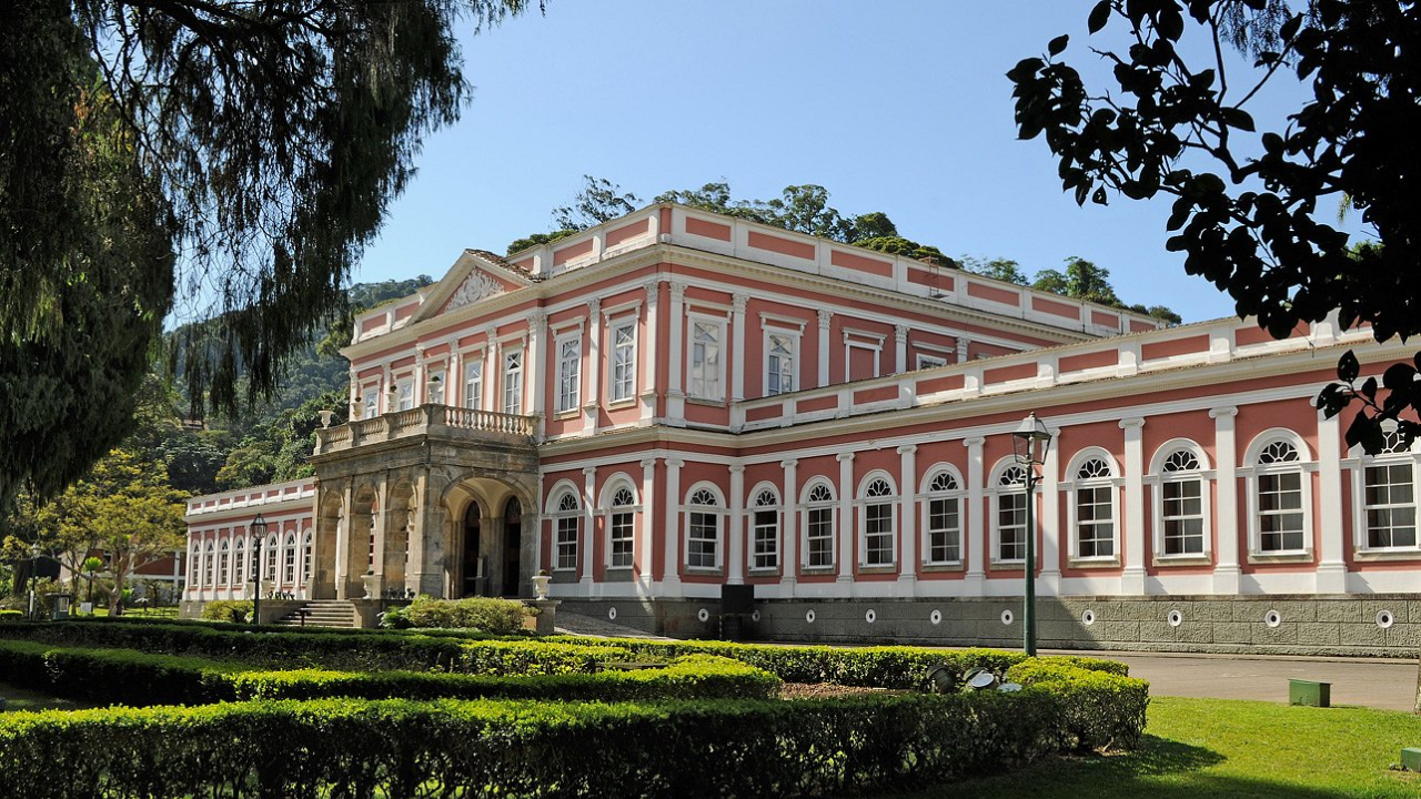 Fachada do Museu Imperial, em Petrópolis, no Rio de Janeiro, com paredes pintadas de vermelho e os arcos de cor branca, e um jardim verde à frente com céu azul claro acima.