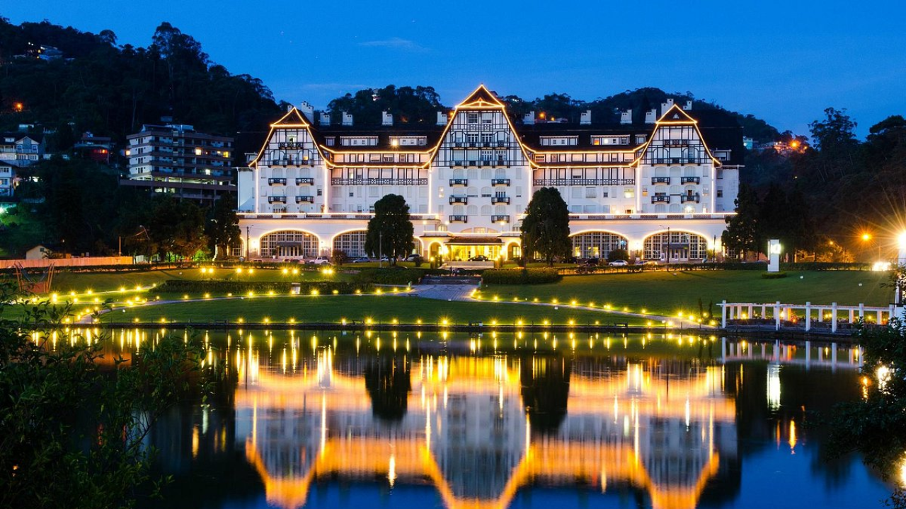 Palácio Quitandinha em Petrópolis, no Rio de Janeiro, ao entardecer, com fachada branca e telhado triangular, típico da arquitetura europeia. À frente há uma grama verde e um lago.