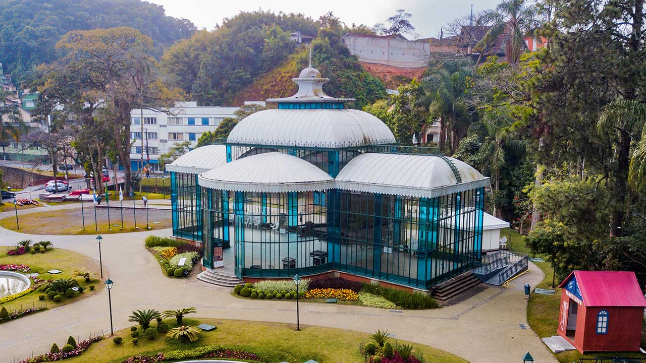 Palácio de Cristal em Petrópolis, no Rio de Janeiro, com arquitetura pré-moldada, paredes de vidro e telhado branco. À frente, há um jardim com flores rosas e amarelas, e o ao fundo há árvores, e o céu está azul claro e sem nuvens.