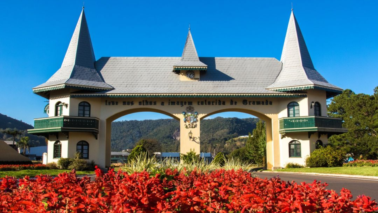 Portal de cor branca com telhado típico da arquitetura europeia em Gramado, no Rio Grande do Sul. À frente há um pequeno jardim de flores vermelhas, uma rua asfaltada e atrás um céu azul claro sem nuvens.