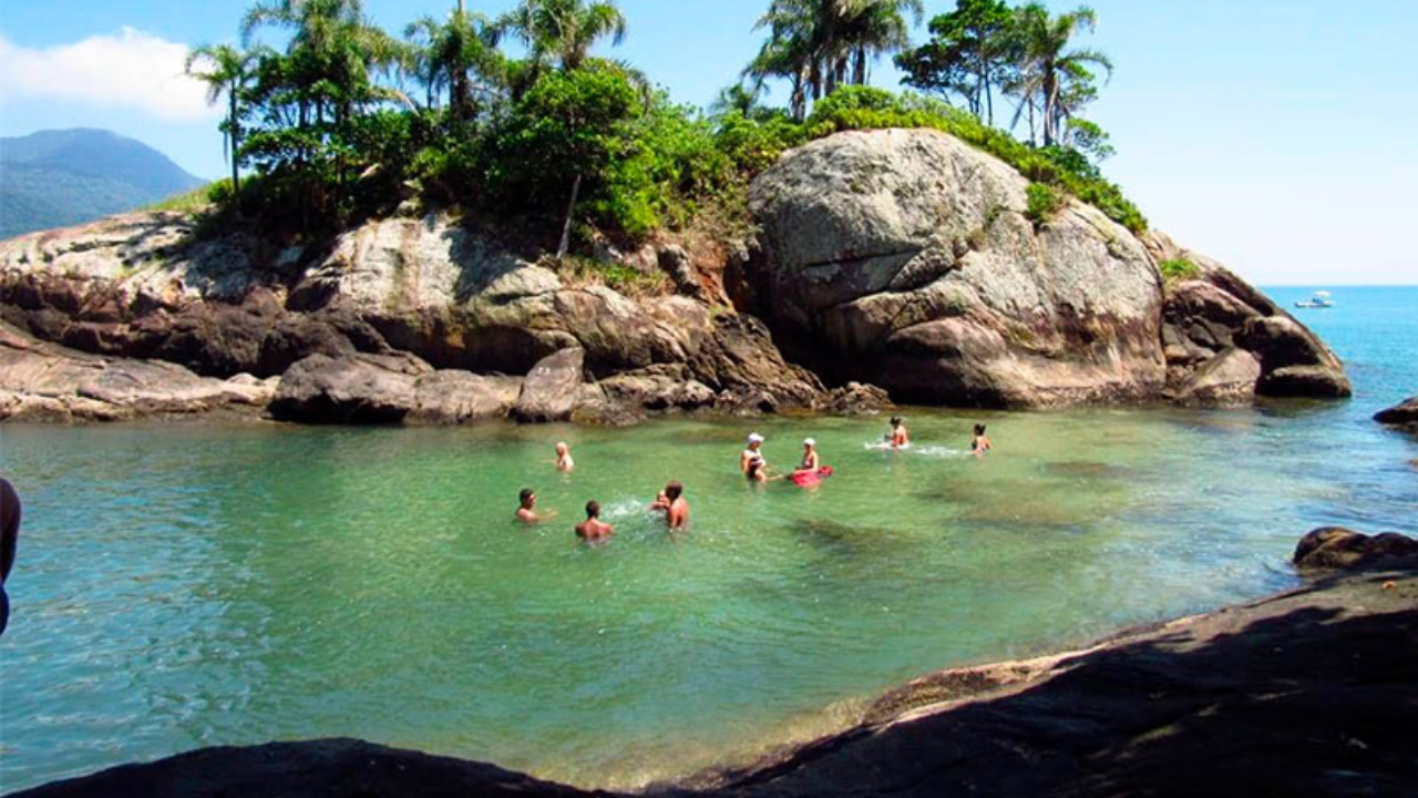 Banhistas em Peruíbe, em São Paulo, em uma pequena área de praia, de água verde cristalina com rochas e árvores ao fundo.
