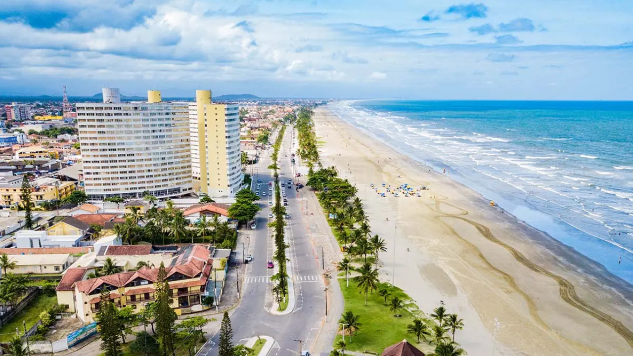 Cidade de Peruíbe, no litoral paulista, com prédios e construções de um lado, uma extensa avenida, um linha verde com grama e coqueiros, e do outro lado, areia e praia, com pequenas ondas se formando. Ao fundo, o céu azul claro, com algumas nuvens. 