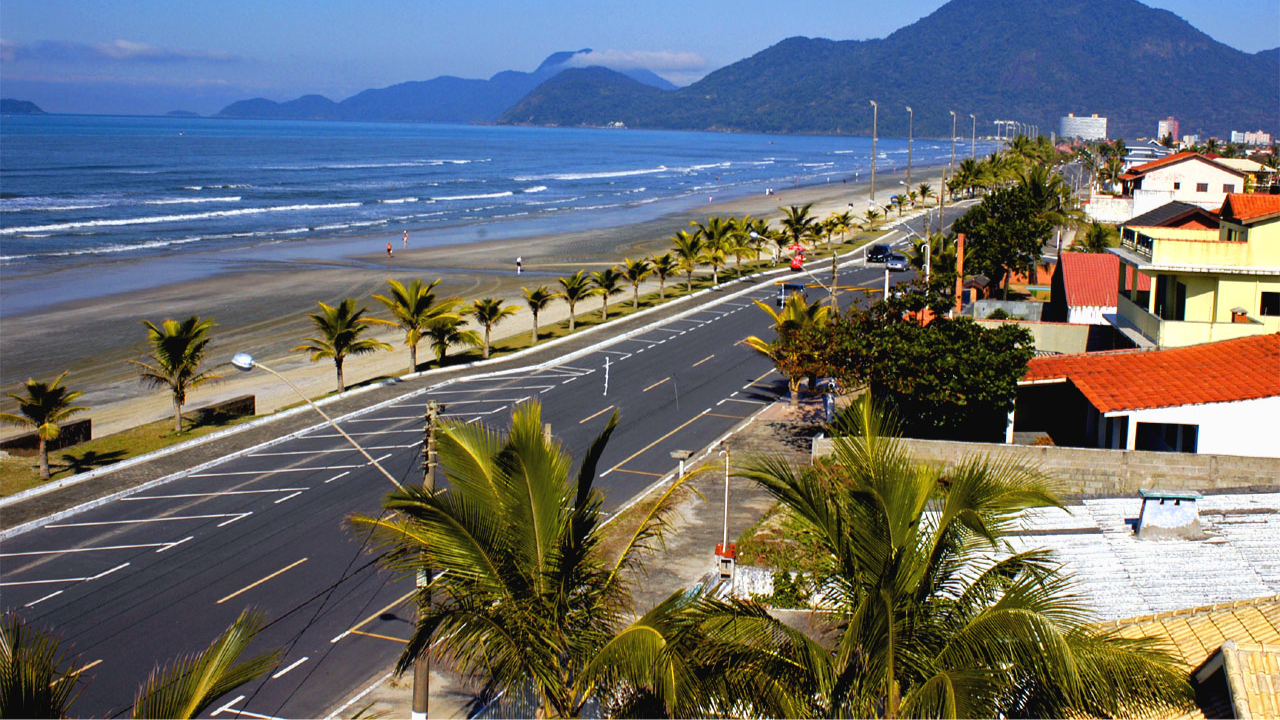 Peruíbe, em São Paulo, em dia de sol, com céu azul claro e sem nuvens, palmeiras ao longo da avenida asfaltada e do outro lado o mar com morros ao fundo.