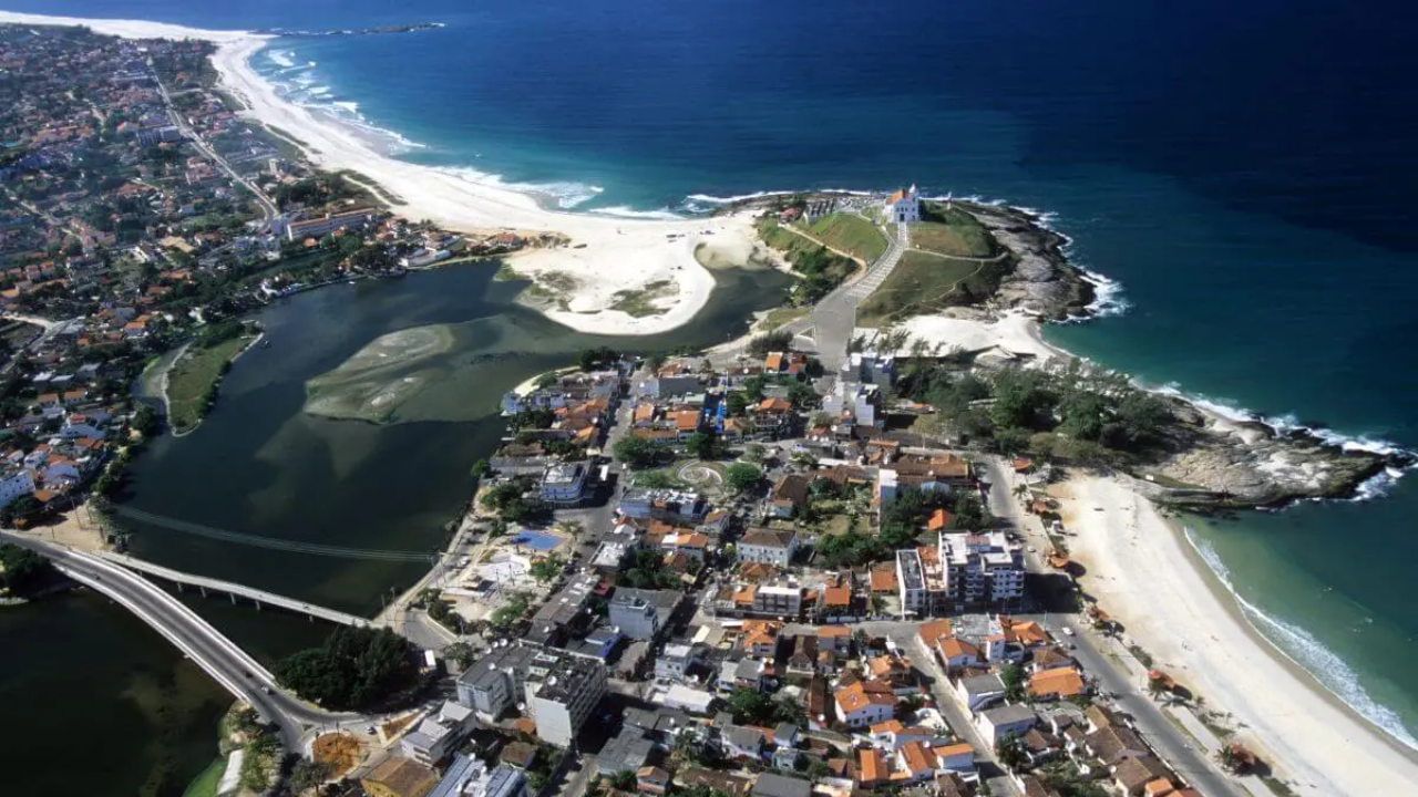 Imagem aérea de Saquarema, no Rio de Janeiro, em que se observa uma ponte cruzando um pequeno rio e conectando os dois lados da cidade, repletos de casas e com o mar à frente. Do outro lado, há praia de areia branca e à frente, um mar que mescla tons de azul. 