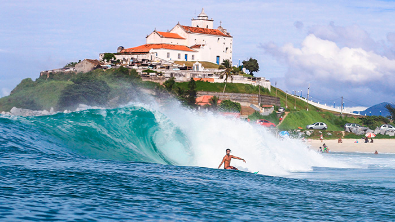 Homem surfando onda em praia de Saquarema, com alguns banhistas no mar e na praia, um casarão ao fundo, algumas árvores e céu azul claro.