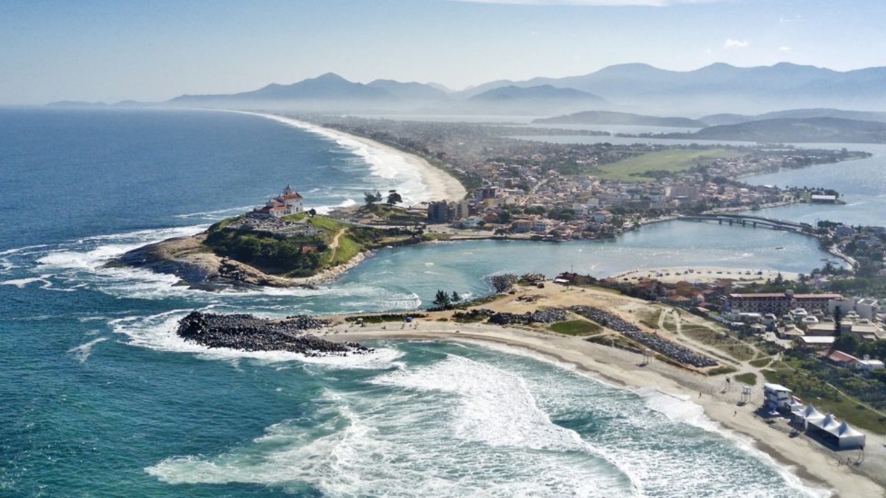 Vista aérea de Saquarema, com as ondas do mar batendo nos rochedos e formando espumas, e alguns morros ao fundo.