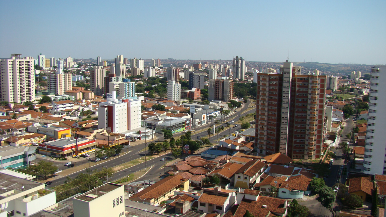 Bauru em dia de sol, com uma avenida comprida em meio às casas e prédios. O céu está claro e limpo, sem nuvens.