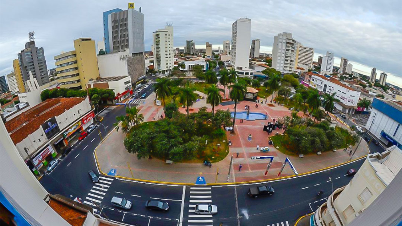 Foto panorâmica de Araçatuba, no interior de São Paulo, em que vemos uma praça no centro da cidade, com árvores, avenida asfaltada em volta com carros passando, e prédios no entorno. O céu está claro com algumas nuvens. 