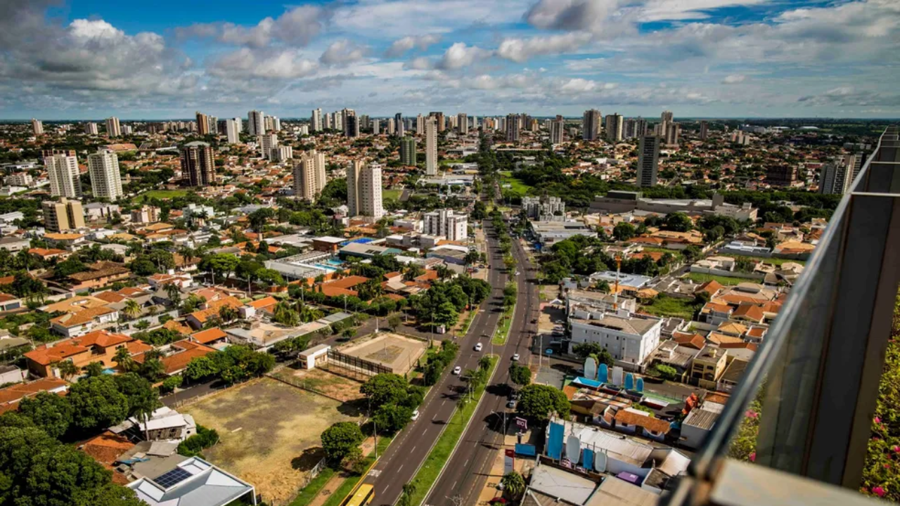 Vista de cima de prédios e casas de Araçatuba, com árvores em meio ao concreto da cidade, em dia claro com algumas nuvens no céu.