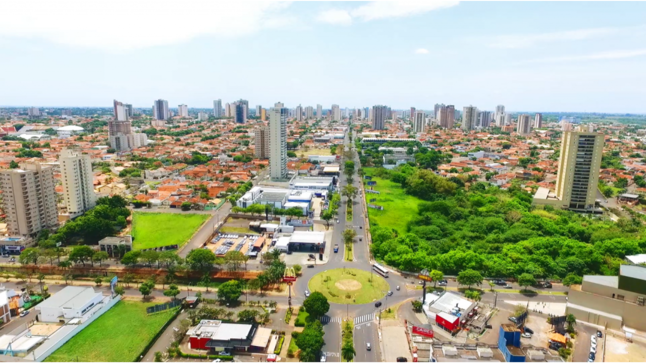 Avenida asfaltada no centro de Araçatuba, em São Paulo, durante o dia, com prédios, casas e estabelecimentos, com uma grande área verde no lado direito contrastando com o concreto da cidade. O céu está claro e com poucas nuvens.