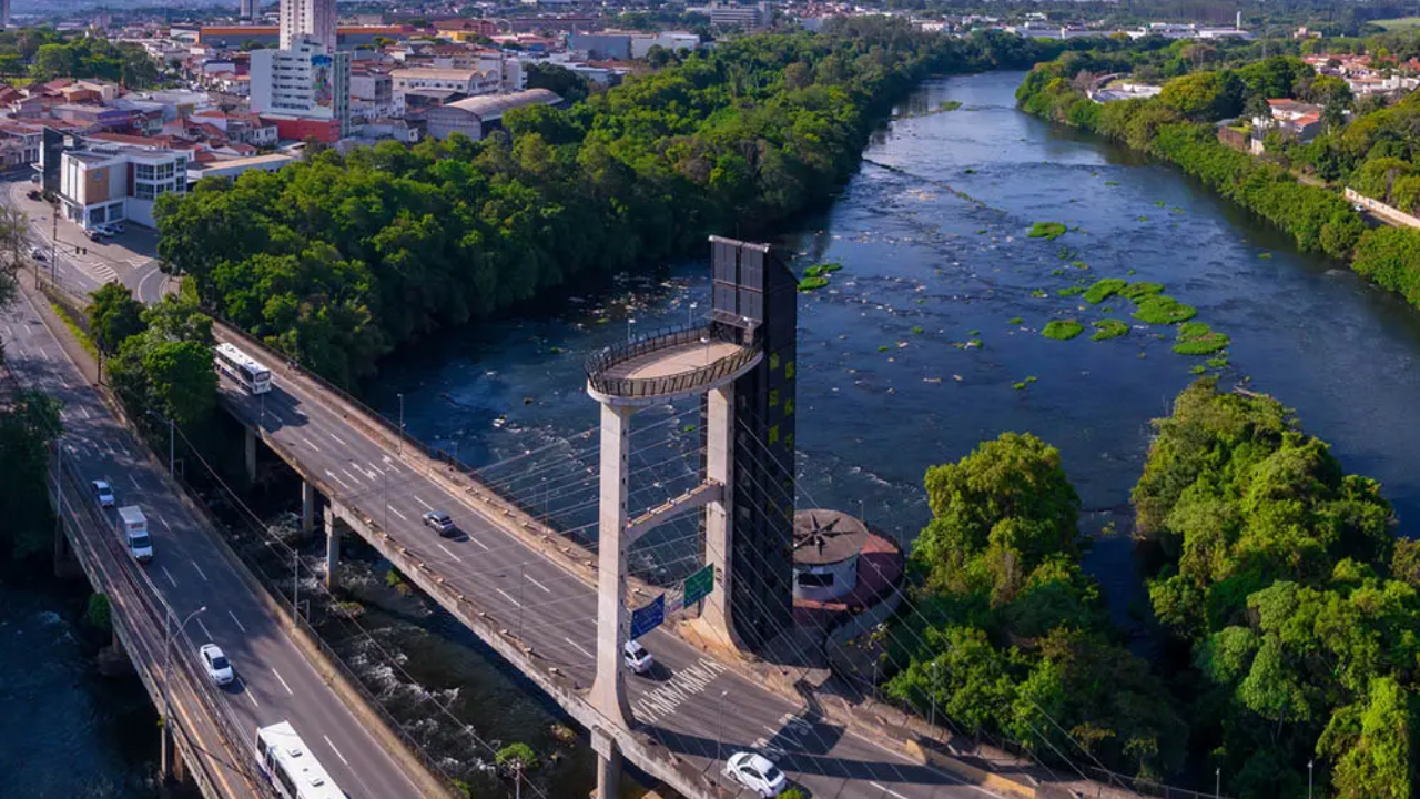 Foto de Piracicaba, no interior de São Paulo, vista de cima, com céu azul claro, sem nuvens, no horizonte, prédios altos, casas, árvores, um rio e uma ponte com carros passando.