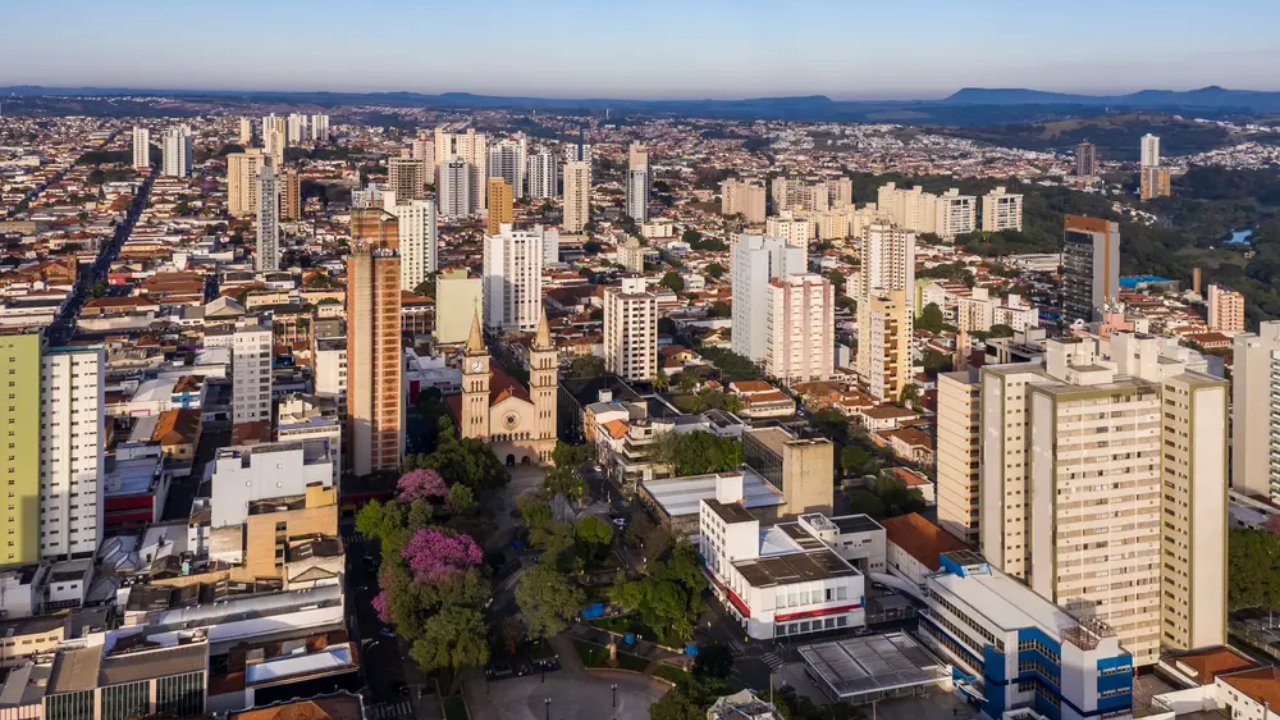 Piracicaba vista de cima, com céu azul claro e limpo, montanhas ao fundo, prédios altos e algumas árvores verdes e roxas mais à frente.
