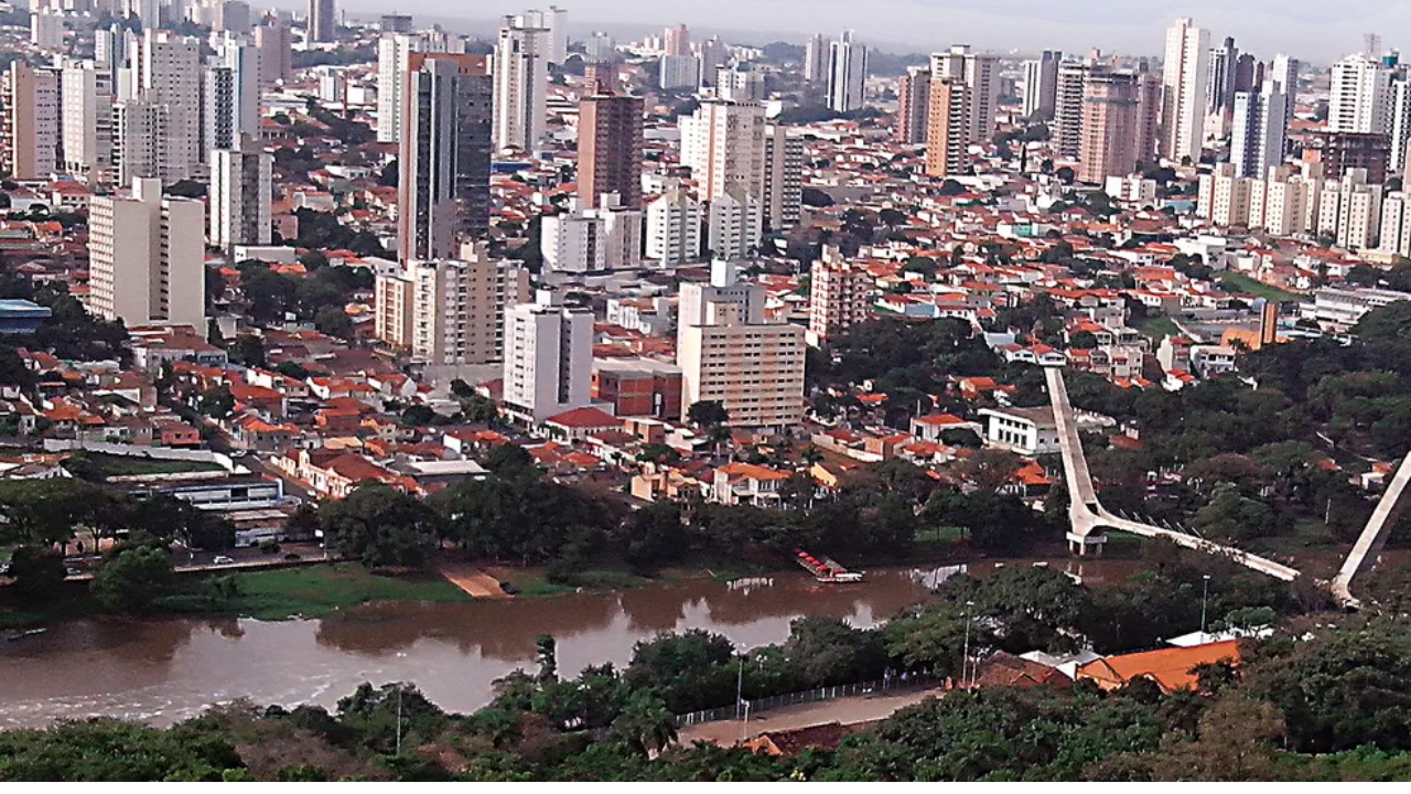 Foto aérea de Piracicaba, com céu azul claro no horizonte, prédios e casas ao fundo e, mais à frente, uma área verde com uma ponte atravessando um rio.