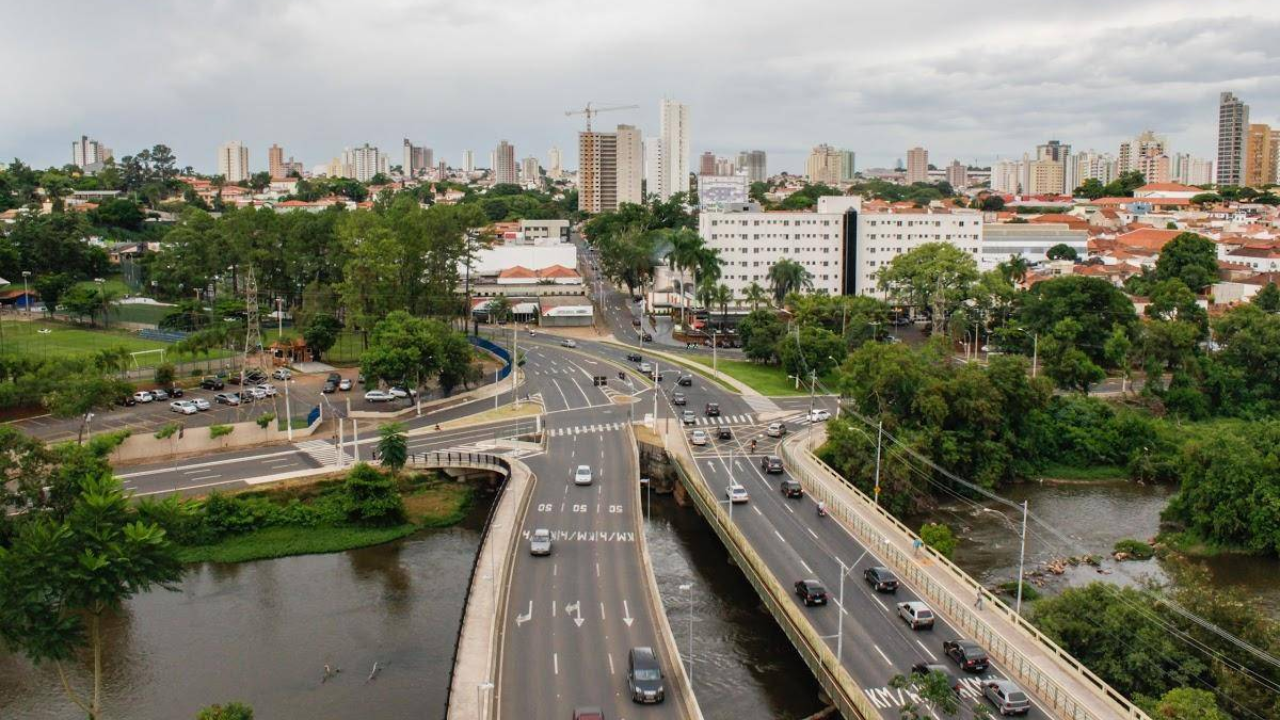 Uma das principais avenidas de Piracicaba, fotografada de cima, com duas pistas paralelas cruzando um rio, com a presença de carros e, entre as construções de concreto da cidade, árvores de copas extensas e esverdeadas. Ao fundo, o céu está claro e com poucas nuvens. 