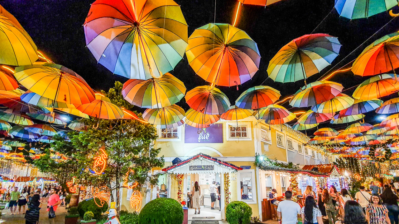 Rua tradicional de Penedo, no interior do Rio de Janeiro, com estabelecimentos iluminados, pessoas andando pelas ruas e, no alto, uma decoração de confetes e guarda-chuvas coloridos.