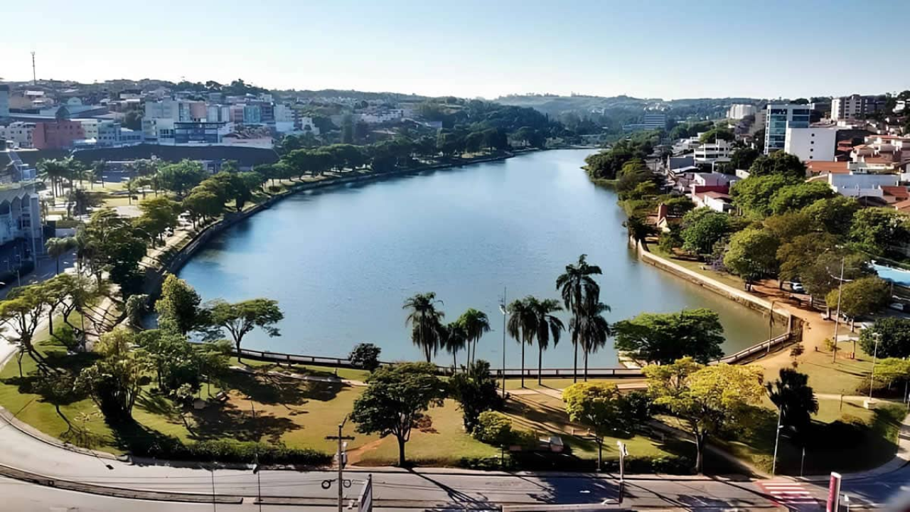 Na foto vemos uma avenida ensolarada, uma pequena faixa verde com grama e Palmeiras, e o rio que corta Bragança Paulista. Ao fundo, temos também alguns prédios.