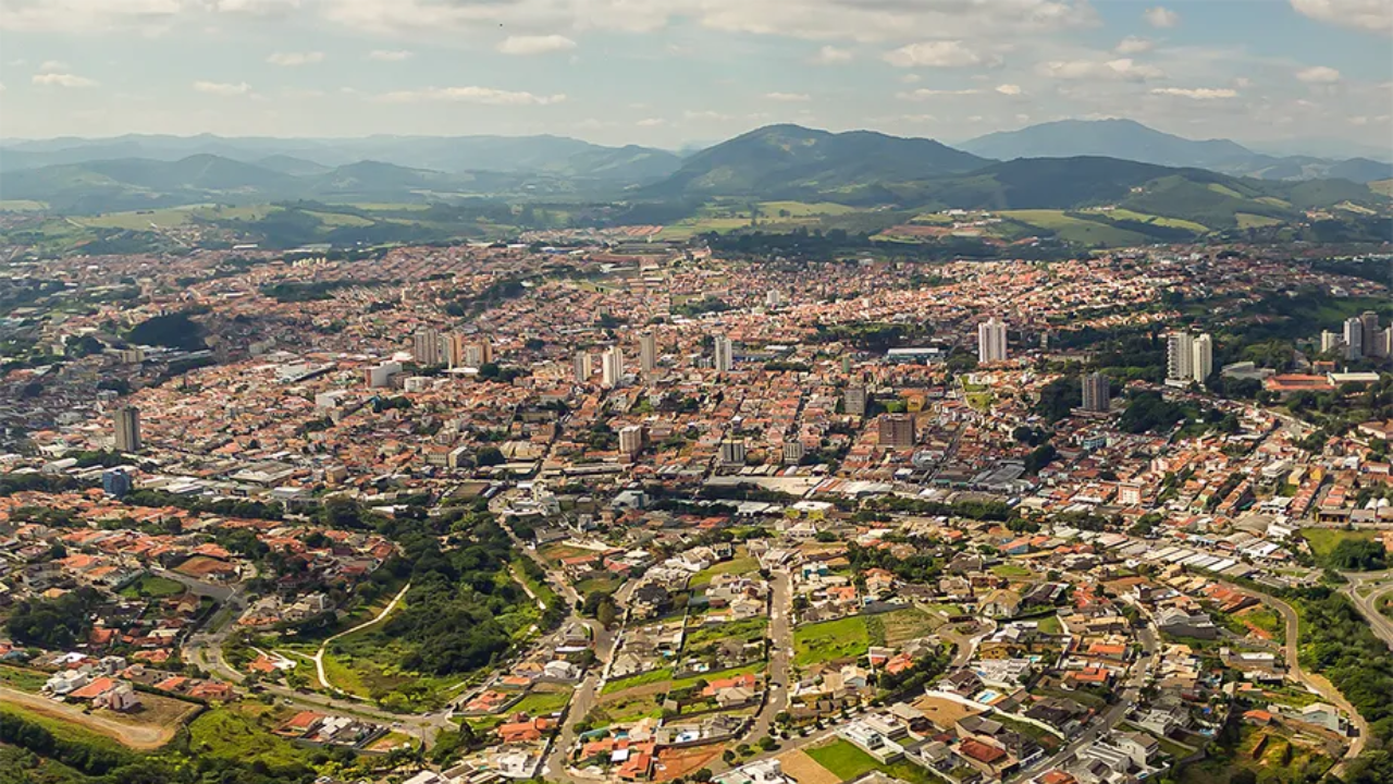 Vista aérea de Bragança Paulista, no interior de São Paulo. Sob o céu azul claro e com poucas nuvens, vemos montanhas ao fundo, casas e prédios logo no centro da imagem e uma pequena área verde logo abaixo.