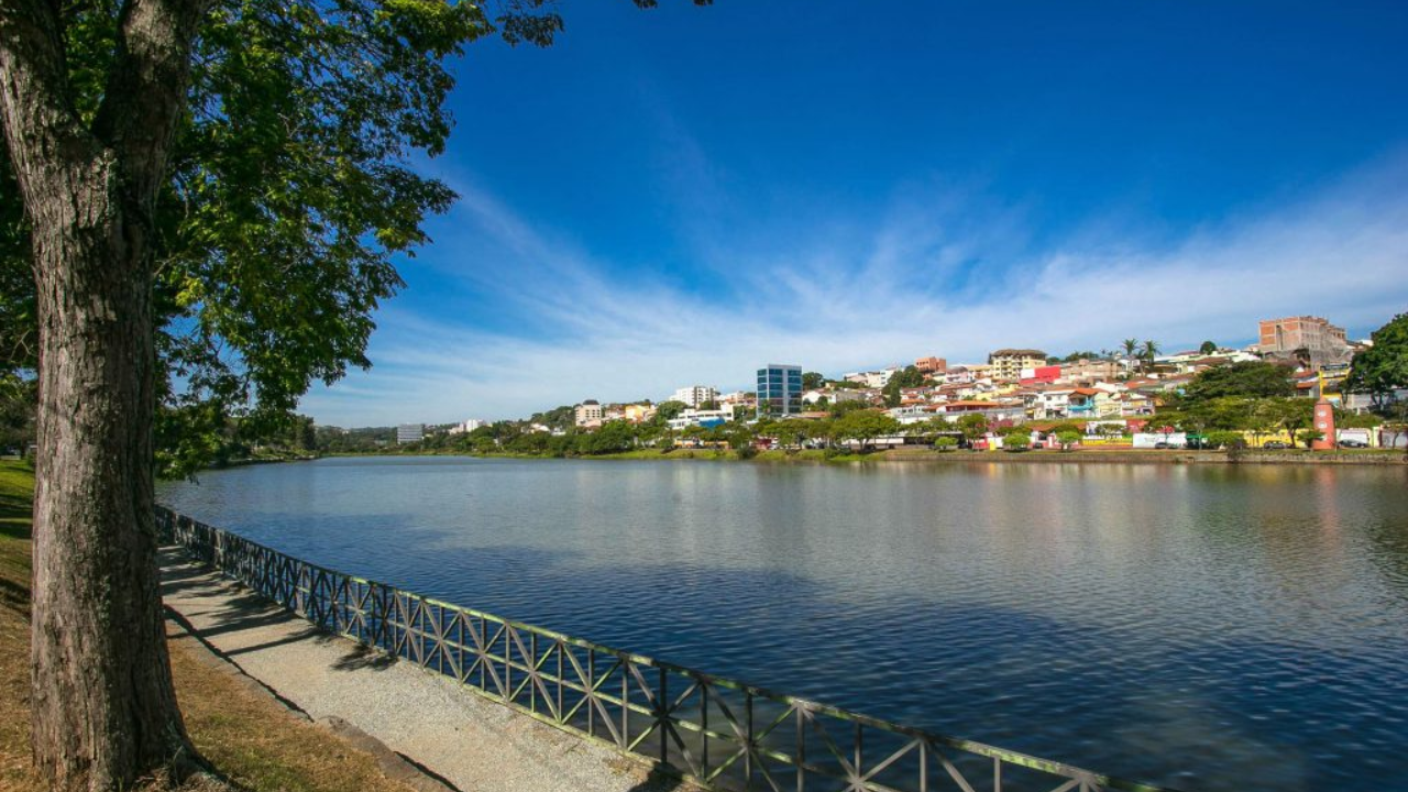 Na foto, vemos o rio que corta a cidade de Bragança Paulista com prédios ao fundo e um céu azul sem nuvens.