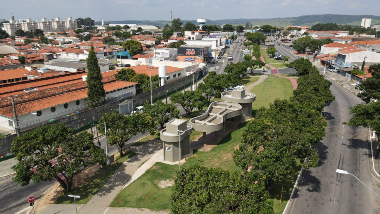 Itu vista de cima durante o dia, com casas ao fundo, uma avenida com carros passando, e uma área verde com árvores e um monumento de concreto.