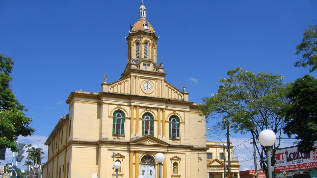 Catedral de Itu, de cor amarela, com céu azul claro e sem nuvens ao fundo, uma passarela espaçosa, postes de iluminação e árvores na frente.