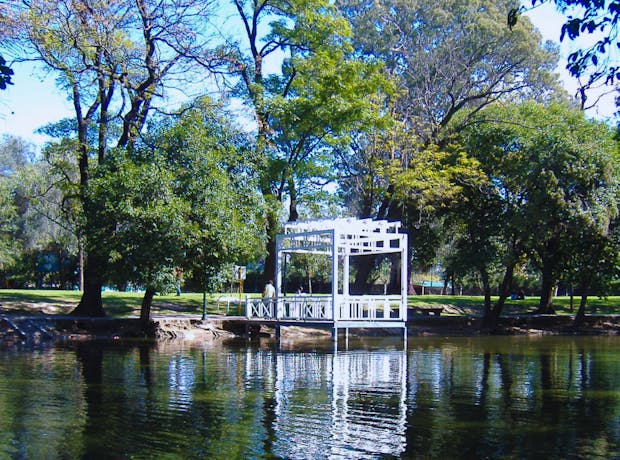 Mirante branco às margens do lago no Parque Sarmiento, em Córdoba, Argentina, rodeado por árvores verdes e refletido na água em um dia ensolarado