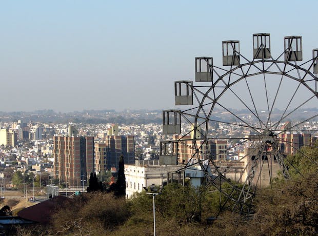 Vista panorâmica da cidade de Córdoba, na Argentina, com prédios residenciais ao fundo e uma roda-gigante metálica à direita, cercada por árvores secas e vegetação, sob um céu claro.
