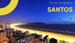 Imagem panorâmica da cidade de Santos ao anoitecer. O céu está azul escuro e as luzes da cidade estão acesas.