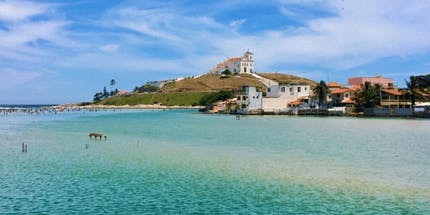 Imagem da Praia da Barrinha na cidade de Saquarema, no Rio de Janeiro. A foto mostra o mar de maré baixa com águas cristalinas e sua costa em uma montanha com algumas casas em um dia ensolarado com o céu azul.