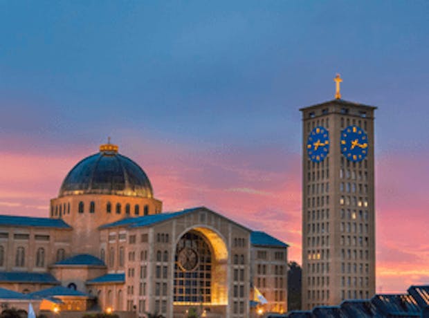 Imagem do Santuário Nacional de Nossa Senhora em Aparecida, São Paulo. A foto mostra a estrutura grandiosa da basílica com suas luzes acesas contra o céu alaranjado e rosado do entardecer.