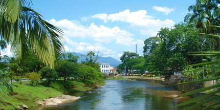 Imagem do Rio Nhundiaquara da cidade de Morretes, no Paraná. A foto mostra a visão central do lago em dia ensolarado, que é cercado por um gramado, árvores e palmeiras.