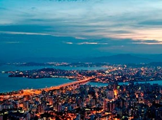 Imagem panorâmica de Florianópolis em Santa Catarina ao entardecer. A imagem mostra diversos prédios com suas luzes acessas, um céu aberto e o mar.