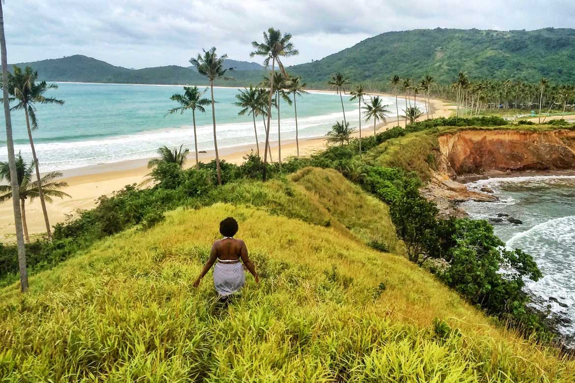 Imagem de uma mulher negra andando em grama alta de costas para uma paisagem praiana, com o oceano ao seu lado esquedo e palmeiras e montanhas à sua frente.