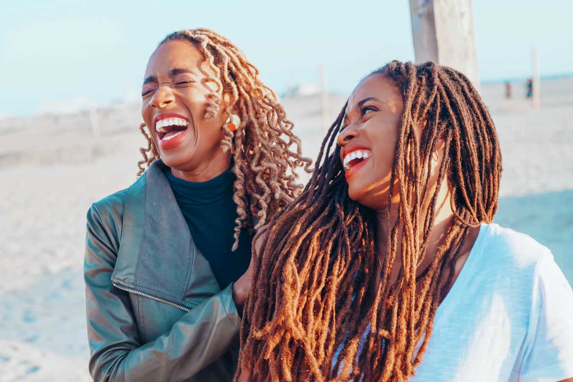 A imagem mostra duas mulheres negras se olhado e dando risada juntas em uma praia ao pôr do sol.