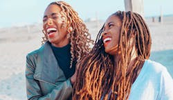 A imagem mostra duas mulheres negras se olhado e dando risada juntas em uma praia ao pôr do sol.